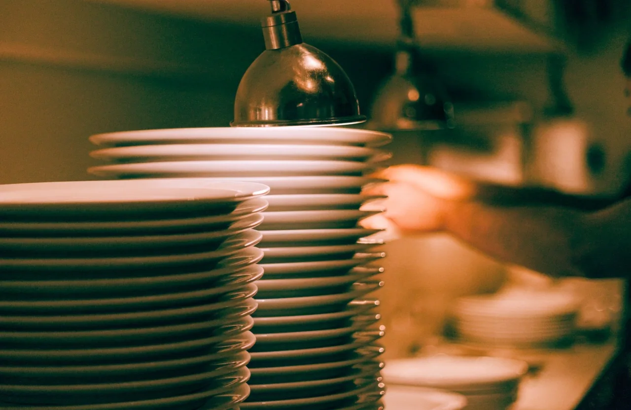 A kitchen pass during dinner service with stacked plates under a heat lamp